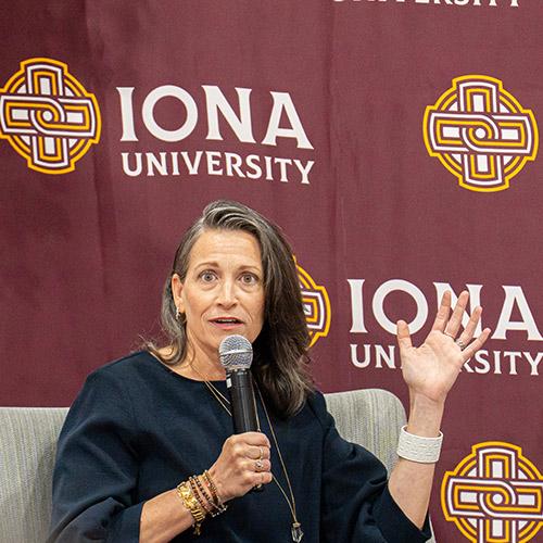 Amy Emmerich ’97 speaks into a microphone while gesturing during her keynote address in front of an Iona University backdrop.