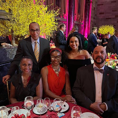 Lucille Bootman smiles alongside a group of formally dressed guests seated around a banquet table at a brightly lit Iona University event decorated with vibrant yellow floral arrangements.
