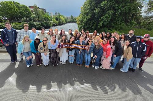 A large group of Iona University study abroad students and faculty pose together on a pedestrian bridge over a river in Ireland while holding up a university scarf.