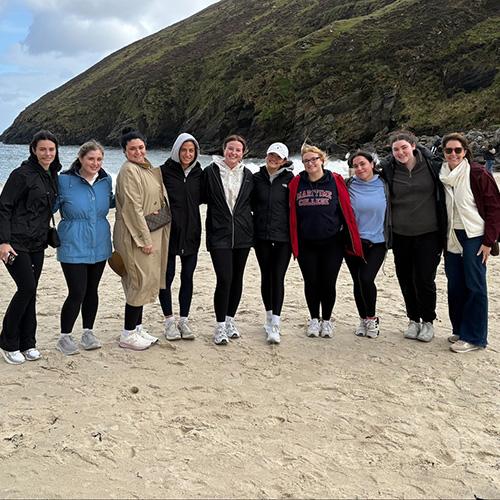 A group of women pose together on a sandy beach in Ireland with a steep green mountain rising in the background.