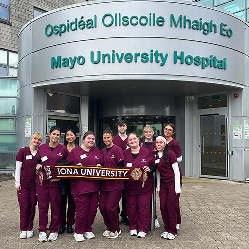 A group of Iona University nursing students wearing maroon scrubs smile and hold up a university scarf in front of the entrance to Mayo University Hospital in Ireland.