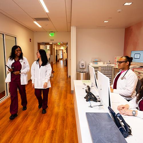 Two healthcare admins walk down the hallway while two others work at the front desk.