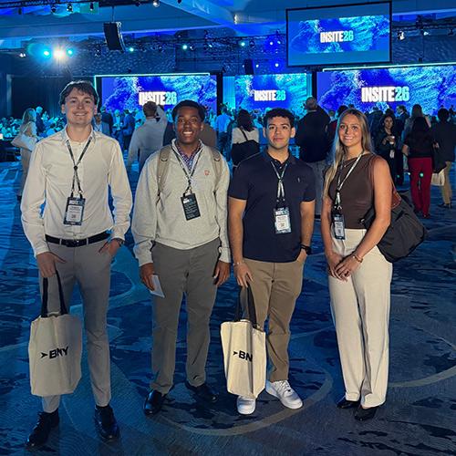 Four Iona University students pose together carrying BNY tote bags inside the convention hall at BNY Insite 2026.