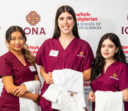 Three nursing students with their white coats in front of an Iona step and repeat.