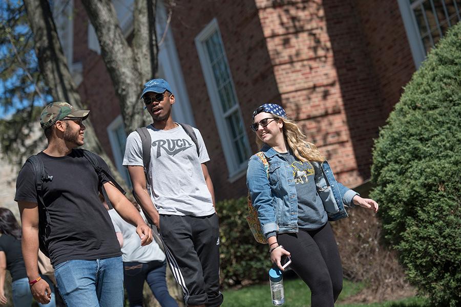 Two male students and one female student walk on campus and smile and talk. They are all stylish and artsy.