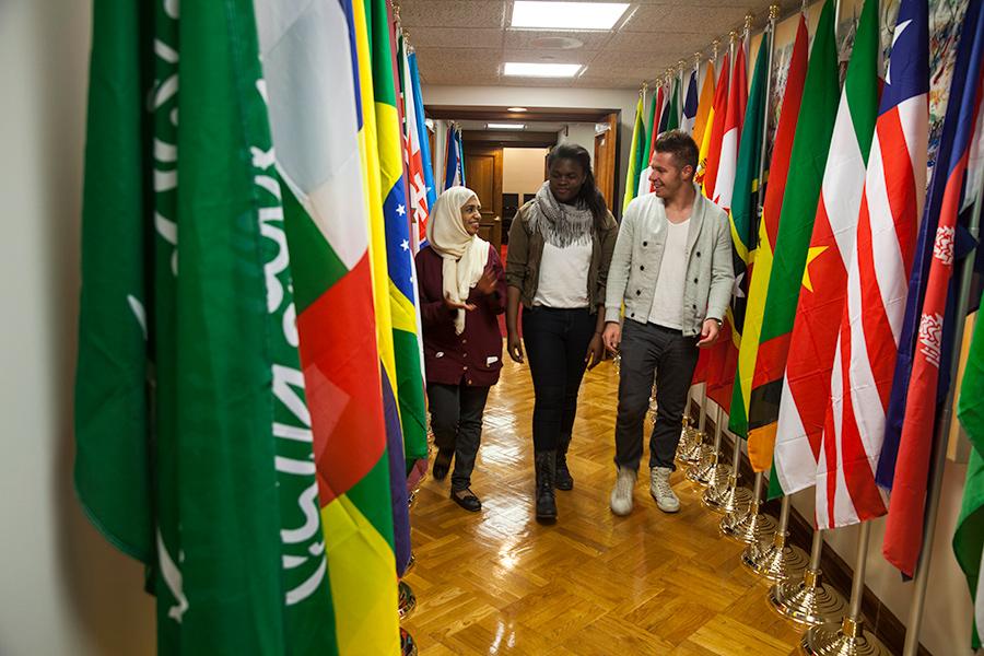 Three international studies students walks down a hallway lined with flags from different countries.