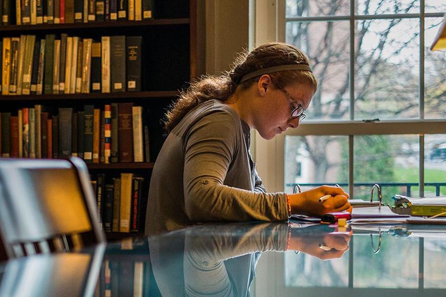 A student sits at a table in the library by the window and writes with pen and paper.