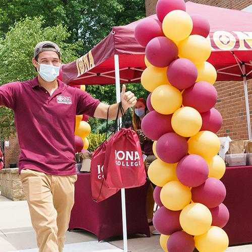 An orientation leader carries tote bags and swag near a table with balloons to welcome new Iona students.